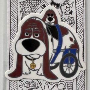 以下が含まれることがあります： 車椅子に乗った2匹の漫画の犬が描かれたステッカー。犬は茶色、白、黒で、「Pope」と書かれた金のタグが付いています。背景には落書きと「BassetStudiosCentral.com」と「@BassetStudios」の文字があります。