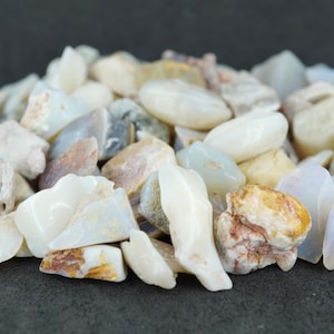 May include: A pile of raw opal stones in various shapes and sizes. The stones display a range of colors, including white, cream, tan, and hints of blue and purple. The opals are rough-cut and appear natural.