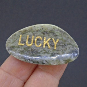 May include: A green and gray stone with the word "LUCKY" in gold lettering. The stone is oval-shaped and has a natural, textured appearance. The gold lettering is centered on the stone.
