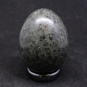 Könnte beinhalten: Ein gesprenkeltes, dunkelgrünes Steinei, hochglanzpoliert. Das Ei wird auf einem kleinen, silberfarbenen Metallständer präsentiert. Die Oberfläche des Eis hat ein gesprenkeltes Aussehen mit dunkleren grünen und schwarzen Flecken.