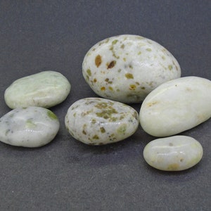 May include: A collection of six smooth, oval-shaped stones. The stones vary in size and have a creamy white base color with green and brown speckles. Some stones have a solid, pale green hue.