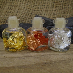 Puede incluir: Tres botellas de vidrio con líquido transparente y escamas metálicas en su interior. Las botellas son de vidrio transparente con un tapón de corcho. Las escamas son doradas, cobrizas y plateadas.
