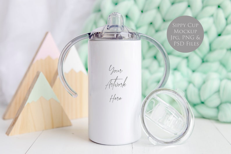 May include: White stainless steel sippy cup with two handles and a clear lid. The cup is on a white surface with wooden mountains in the background. The text "Your Artwork Here" is printed on the cup.