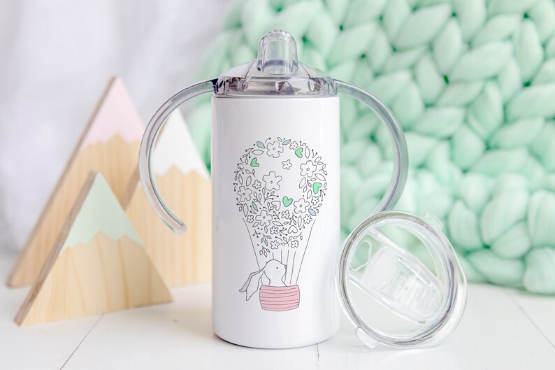 May include: White stainless steel toddler sippy cup with a clear lid and two handles. The cup features a design of a hot air balloon with a bunny rabbit inside and a floral pattern around the balloon.