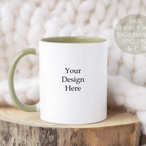May include: White mug with a sage green handle and interior, resting on a wooden slice. The mug has the text "Your Design Here" printed on the front. The background is a soft, white, textured fabric. The mug is a mockup.