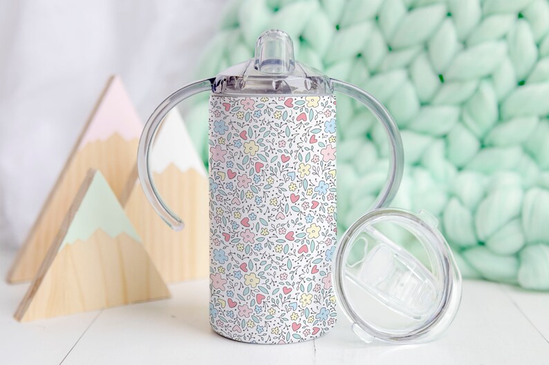 May include: A white stainless steel tumbler with a floral pattern in pink, blue, and yellow. The tumbler has two handles and a clear lid with a straw hole.