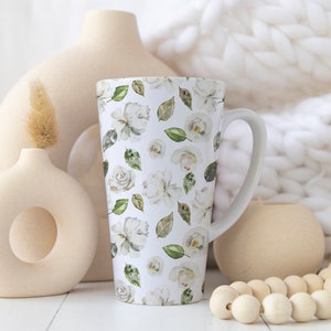 May include: Tall white ceramic mug with a floral pattern of white roses and green leaves. The mug has a curved handle and is surrounded by neutral-toned vases and a wooden bead garland. The background includes a white, textured blanket.