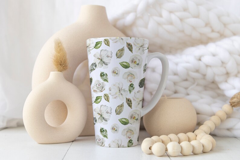 May include: Tall white ceramic mug with a floral pattern of white roses and green leaves. The mug has a curved handle and is surrounded by neutral-toned vases and a wooden bead garland. The background includes a white, textured blanket.