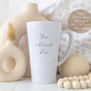 May include: A tall white latte mug mockup with the text "Your Artwork Here". The mug is displayed with beige vases, a decorative wooden bead garland, and a soft white blanket in the background. The image also includes a circular graphic with the text "TALL LATTE MUG MOCKUP JPG, PSD AND PNG FILES".