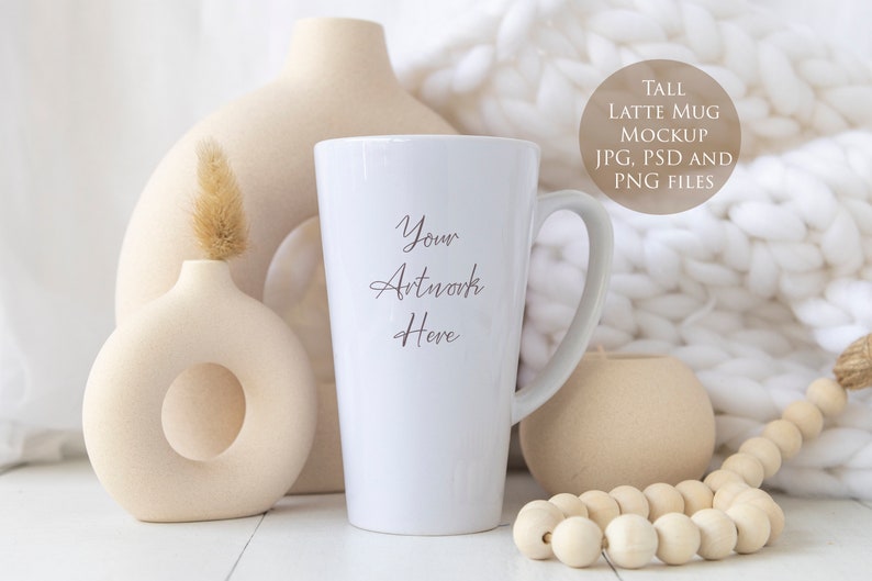 May include: A tall white latte mug mockup with the text "Your Artwork Here". The mug is displayed with beige vases, a decorative wooden bead garland, and a soft white blanket in the background. The image also includes a circular graphic with the text "TALL LATTE MUG MOCKUP JPG, PSD AND PNG FILES".