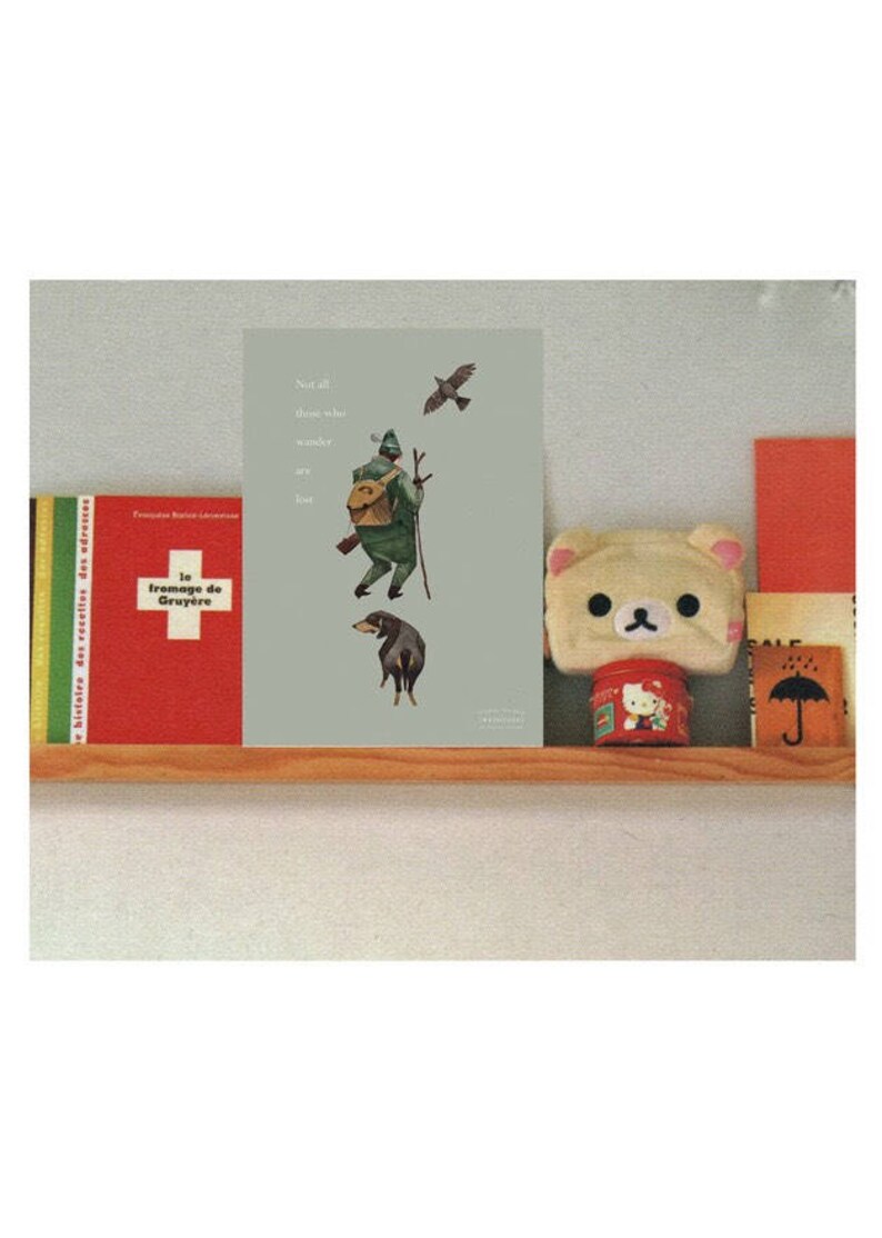 May include: A framed print with a light green background featuring a watercolor illustration of a person in green clothing with a backpack, walking with a stick and a dog following behind. The text "Not all those who wander are lost" is printed in white on the top left corner of the print.
