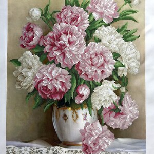 Pode incluir: Um buquê de peónias rosa e brancas num vaso branco e dourado com base verde. O vaso está sobre uma toalha de renda branca. O fundo é de cor castanho claro.