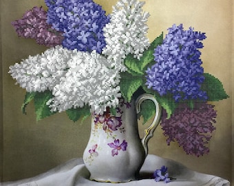 May include: A bouquet of purple, white, and lavender lilac flowers in a white porcelain jug with a floral pattern. The jug is sitting on a white tablecloth with lace trim.