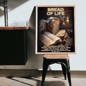 Op de afbeelding: Een ingelijste print met de tekst "BREAD OF LIFE" in een gloeiend lettertype. De afbeelding toont een brood, een open boek, een kaars en een kelk. De tekst hieronder luidt "BUT HE ANSWERED AND SAID, IT IS WRITTEN..." Mattheüs 4:4 KJV.