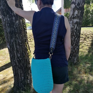K&ouml;nnte beinhalten: T&uuml;rkisfarbene H&auml;keltasche mit Schulterriemen und Muster. Die Tasche hat goldfarbene Beschl&auml;ge und einen Riemen mit geometrischem Design in Blau, Lila und Wei&szlig;. Die Tasche wird &uuml;ber der Schulter getragen. Die Tasche ist einfarbig.