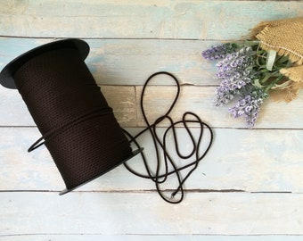 May include: A spool of dark brown cord sits next to a length of the same cord, unraveled. The cord is a solid color and appears to be made of a woven material. The background includes lavender sprigs in a burlap wrap.