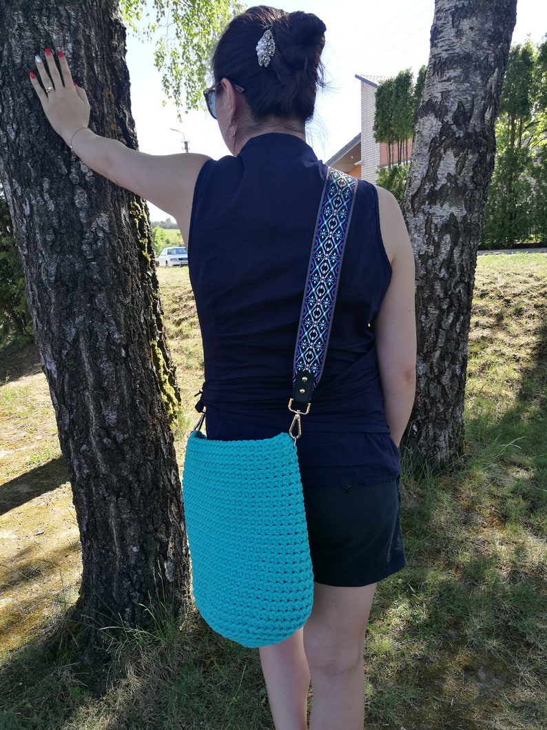 K&ouml;nnte beinhalten: T&uuml;rkisfarbene H&auml;keltasche mit Schulterriemen mit Muster. Die Tasche ist einfarbig und hat eine zylindrische Form. Der Riemen ist schwarz mit einem blauen und wei&szlig;en geometrischen Design. Die Tasche h&auml;ngt an einem goldfarbenen Verschluss.