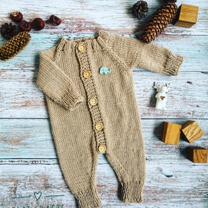 Peut inclure: Une grenouillère bébé en tricot beige avec une fermeture boutonnée sur le devant. La grenouillère a une petite application en forme d'éléphant sur la poitrine. La grenouillère est photographiée sur un fond en bois bleu clair avec des pommes de pin et des blocs de bois.