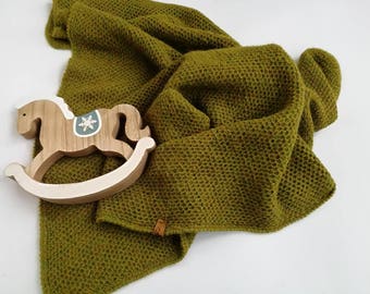 Pode incluir: Uma manta de malha verde com um padrão texturizado, dobrada e drapeada sobre um cavalo de balanço de madeira com um desenho de floco de neve branco.