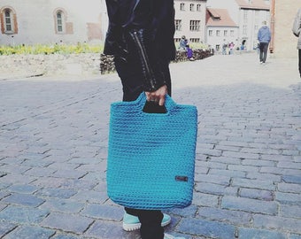 May include: A large, turquoise blue crocheted tote bag with two handles. The bag has a rectangular shape and a small, rectangular brown tag on the front. The bag is being held by a person wearing black pants, a black jacket, and turquoise shoes.