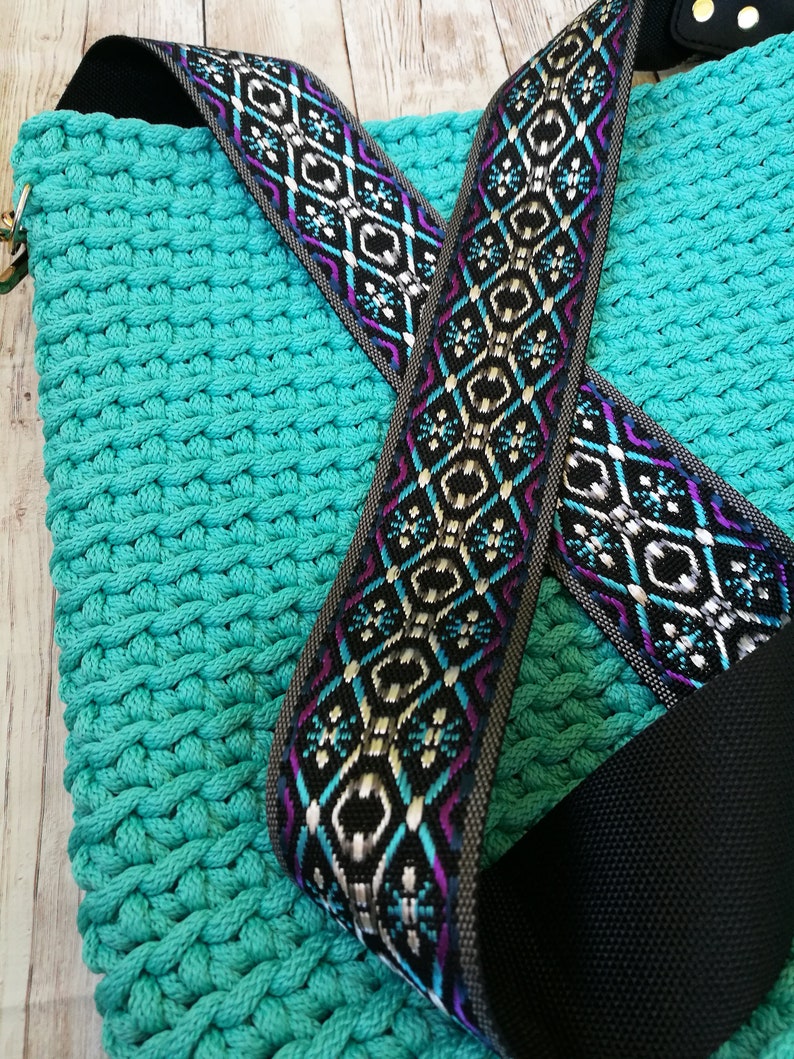 K&ouml;nnte beinhalten: Eine t&uuml;rkisfarbene Makramee-Tasche mit einem gemusterten Riemen. Der Riemen hat einen schwarzen Hintergrund mit geometrischen Designs in Wei&szlig;, Lila und T&uuml;rkis. Die Tasche besteht aus dickem, gewebtem Material.