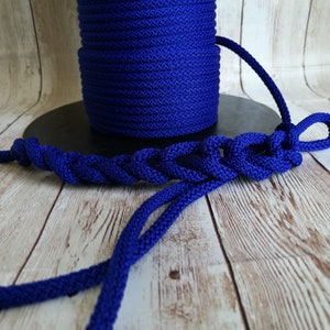 May include: A spool of vibrant blue rope sits next to a length of braided rope. The braided section is looped and knotted, showcasing the rope's texture and color. The background is a light wood grain.