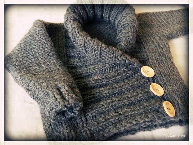 May include: A gray knitted sweater with a large, folded collar and three wooden buttons. The sweater has a textured knit pattern and is likely made of wool or a similar material. The sleeves are visible, and the garment appears to be handmade.