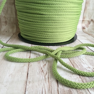 May include: A spool of light green, braided cord sits on a black base, with a length of the same cord in the foreground. The cord is a solid, pale green color, and the background is a light wood grain.