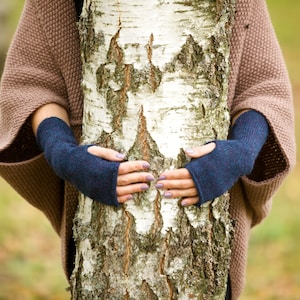 May include: A person wearing a brown knitted cardigan with blue fingerless gloves, holding their hands around a birch tree trunk.