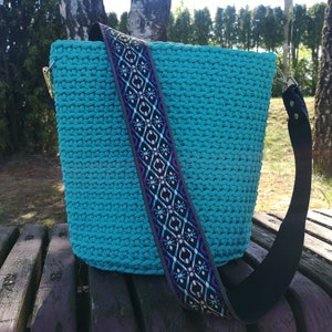 K&ouml;nnte beinhalten: Eine t&uuml;rkisfarbene H&auml;keltasche mit einem gemusterten Riemen. Der Riemen hat ein geometrisches Design in Schwarz, Lila und Wei&szlig;. Die Tasche hat goldfarbene Beschl&auml;ge und einen schwarzen Riemen.
