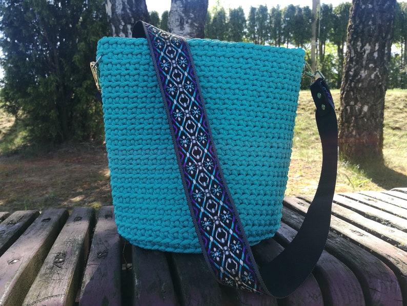 K&ouml;nnte beinhalten: Eine t&uuml;rkisfarbene H&auml;keltasche mit einem gemusterten Riemen. Der Riemen hat ein geometrisches Design in Schwarz, Lila und Wei&szlig;. Die Tasche hat goldfarbene Beschl&auml;ge und einen schwarzen Riemen.
