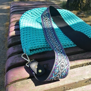 K&ouml;nnte beinhalten: Eine t&uuml;rkisfarbene Stricktasche mit dekorativem Riemen. Der Riemen hat eine schwarze R&uuml;ckseite mit einem Muster in Wei&szlig;, Lila und Blau. Die Tasche hat einen silbernen Verschluss und liegt auf einer Holzbank.
