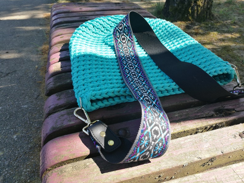 K&ouml;nnte beinhalten: Eine t&uuml;rkisfarbene Stricktasche mit dekorativem Riemen. Der Riemen hat eine schwarze R&uuml;ckseite mit einem Muster in Wei&szlig;, Lila und Blau. Die Tasche hat einen silbernen Verschluss und liegt auf einer Holzbank.