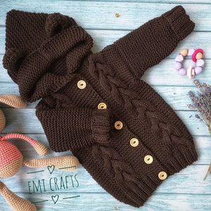 May include: A brown knitted baby cardigan with a hood and button closure. The cardigan has a cable knit pattern and is laid out on a blue wooden surface.