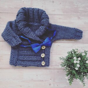 May include: A navy blue knitted baby sweater with a shawl collar and a bright blue bow tie. The sweater has three wooden buttons and is displayed on a light wood surface. A small green plant with white flowers is in the background.