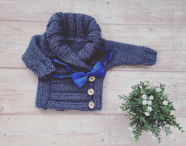 May include: A navy blue knitted baby sweater with a shawl collar and a bright blue bow tie. The sweater has three wooden buttons and is displayed on a light wood surface. A small green plant with white flowers is in the background.