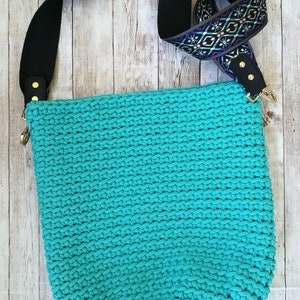 K&ouml;nnte beinhalten: Eine t&uuml;rkisfarbene H&auml;keltasche mit einem schwarzen Schulterriemen mit Muster. Der Riemen hat ein geometrisches Design in Lila-, T&uuml;rkis- und Wei&szlig;t&ouml;nen. Die Tasche hat goldfarbene Hardware und steht vor einem hellen Holzhintergrund.