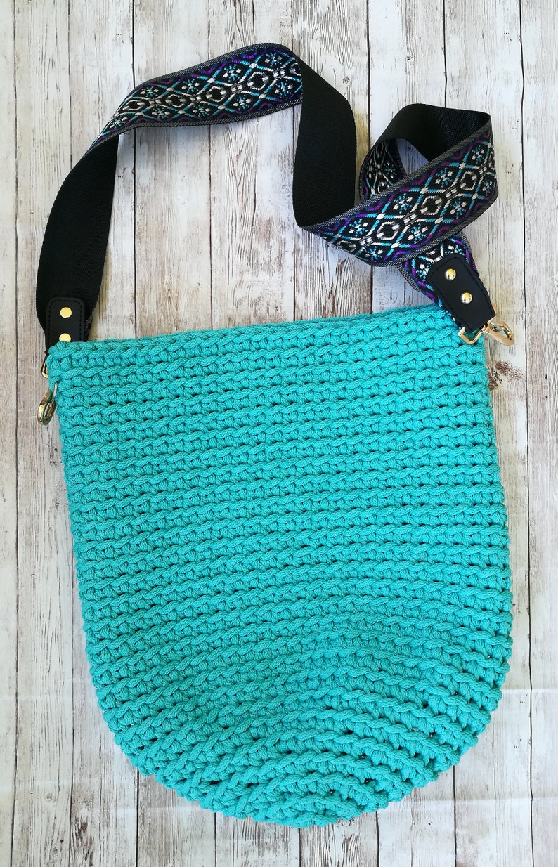 K&ouml;nnte beinhalten: Eine t&uuml;rkisfarbene H&auml;keltasche mit einem schwarzen Schulterriemen mit Muster. Der Riemen hat ein geometrisches Design in Lila-, T&uuml;rkis- und Wei&szlig;t&ouml;nen. Die Tasche hat goldfarbene Hardware und steht vor einem hellen Holzhintergrund.
