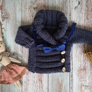 May include: A navy blue knitted baby sweater with a hood and wooden buttons. A blue bow tie is attached. A stuffed bunny wearing a pink skirt is to the left of the sweater. Dried wheat is to the right.