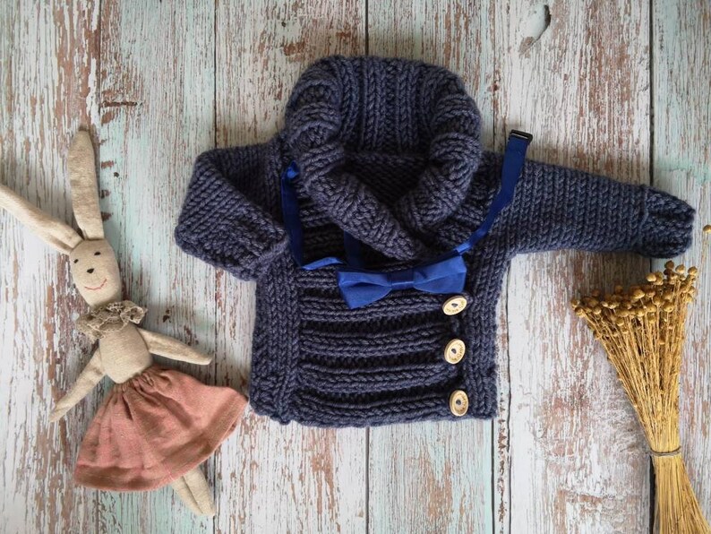 May include: A navy blue knitted baby sweater with a hood and wooden buttons. A blue bow tie is attached. A stuffed bunny wearing a pink skirt is to the left of the sweater. Dried wheat is to the right.