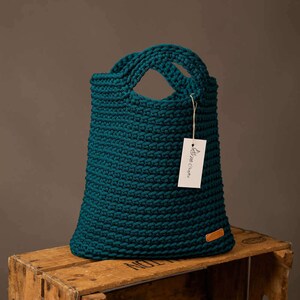 Pode incluir: Uma sacola tote de croch&ecirc; verde-azulado com duas al&ccedil;as. A sacola tem formato retangular e uma etiqueta que diz "Brim Crafts". A sacola est&aacute; em cima de uma caixa de madeira.