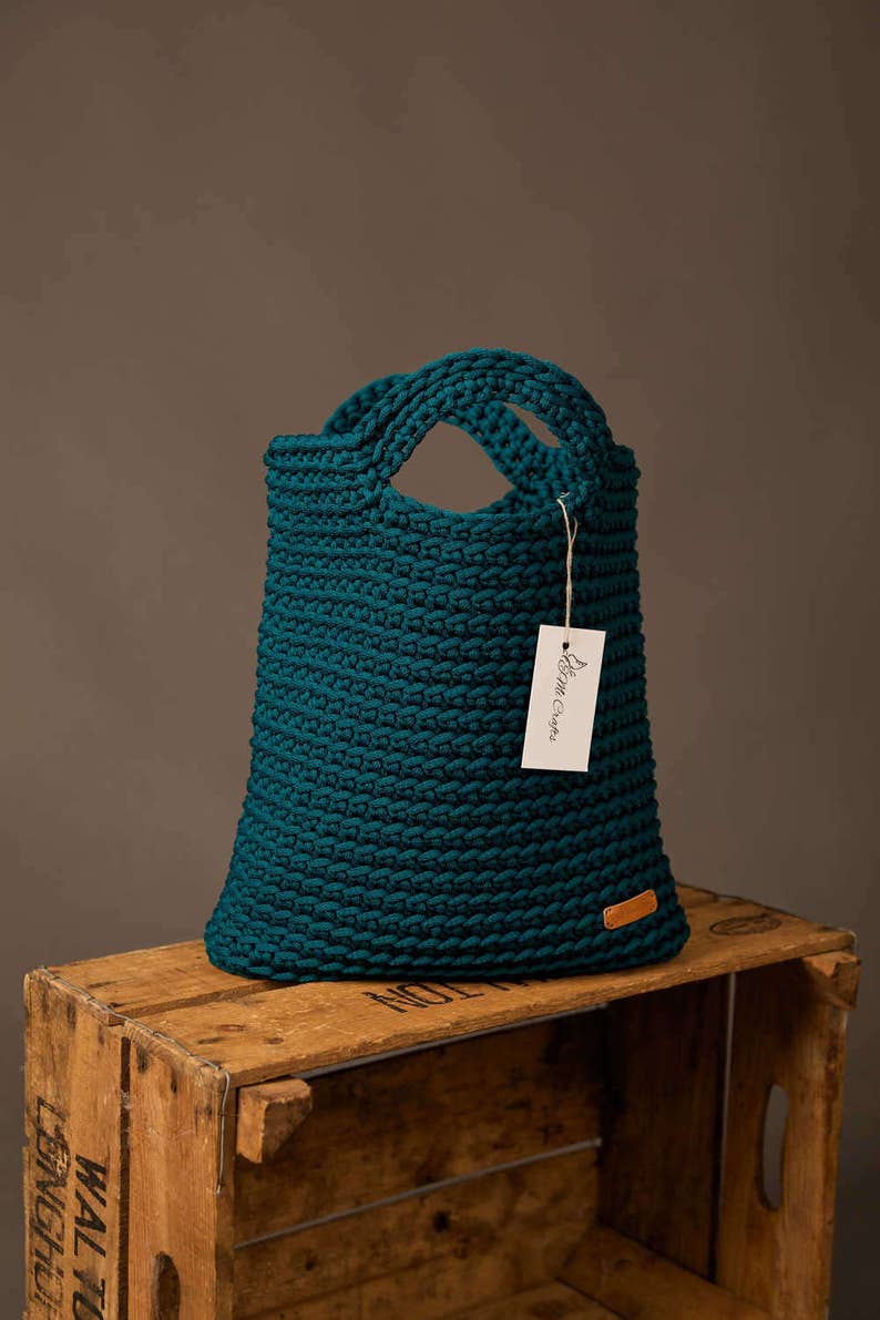 Pode incluir: Uma sacola tote de croch&ecirc; verde-azulado com duas al&ccedil;as. A sacola tem formato retangular e uma etiqueta que diz "Brim Crafts". A sacola est&aacute; em cima de uma caixa de madeira.