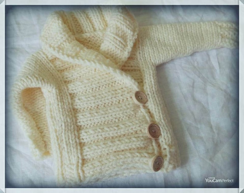 May include: Cream-colored, hand-knitted baby cardigan with a hood. The sweater features a ribbed texture, wooden buttons, and long sleeves. The garment is made of soft yarn, perfect for keeping a baby warm.