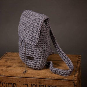 Könnte beinhalten: Ein grauer Häkelrucksack mit Klappenverschluss und verstellbaren Trägern. Der Rucksack hat eine kleine Metallkette, die an der Klappe befestigt ist.