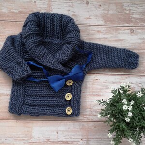 May include: A navy blue knitted baby sweater with a hood and a matching bow tie. The sweater has three wooden buttons and is displayed on a wooden surface. A small plant with white flowers is in the bottom right corner.
