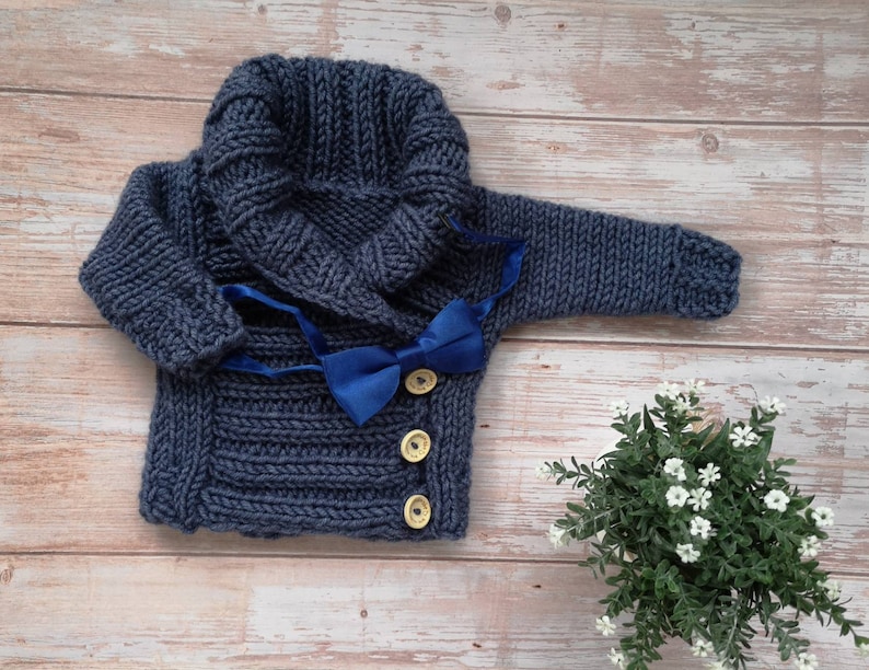 May include: A navy blue knitted baby sweater with a hood and a matching bow tie. The sweater has three wooden buttons and is displayed on a wooden surface. A small plant with white flowers is in the bottom right corner.