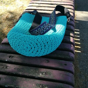 K&ouml;nnte beinhalten: Eine t&uuml;rkisfarbene H&auml;keltasche mit gemustertem Riemen liegt auf einer Holzbank. Der Riemen der Tasche ist schwarz mit einem dekorativen Muster in Lila, Blau und Wei&szlig;. Die Tasche ist rund und hat ein dickes, strukturiertes Aussehen.