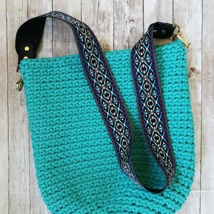 K&ouml;nnte beinhalten: Eine t&uuml;rkisfarbene H&auml;keltasche mit gemustertem Riemen. Der Riemen hat ein geometrisches Design in Schwarz, Wei&szlig;, Lila und Blau. Die Tasche hat ein schwarzes Schulterpolster aus Leder und goldfarbene Beschl&auml;ge.