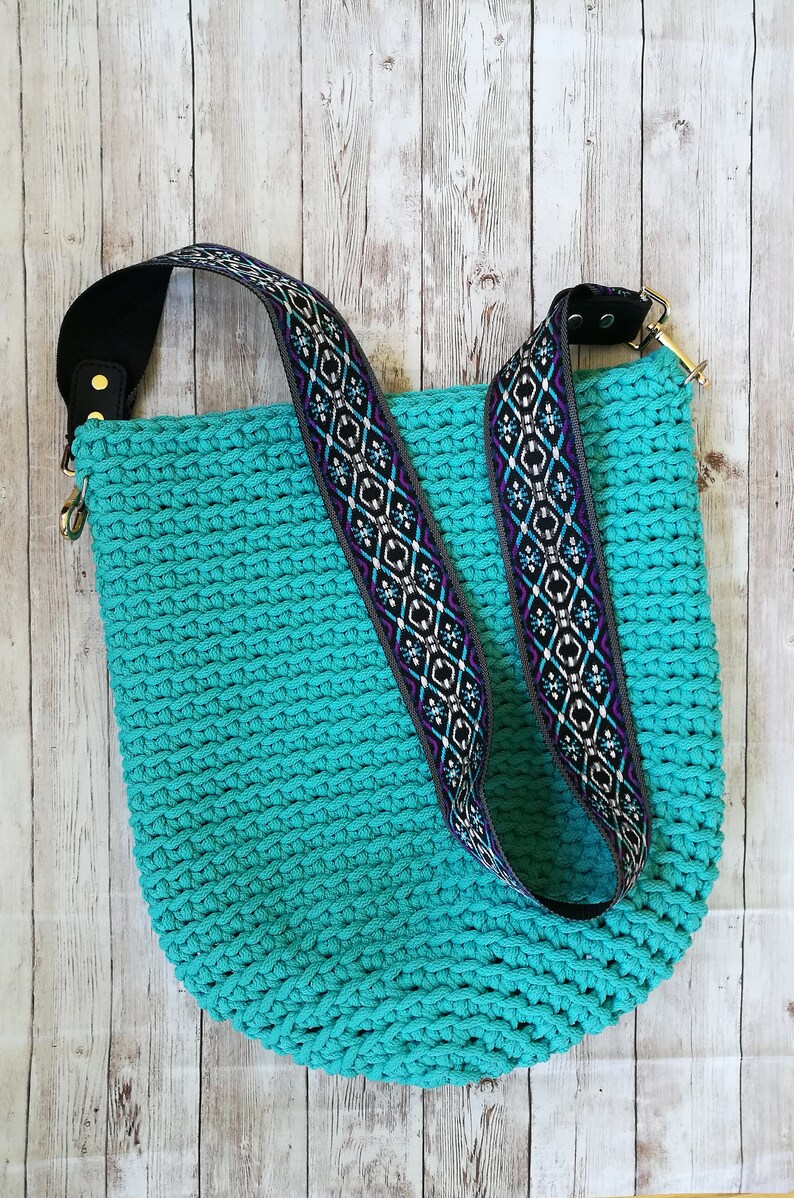 K&ouml;nnte beinhalten: Eine t&uuml;rkisfarbene H&auml;keltasche mit gemustertem Riemen. Der Riemen hat ein geometrisches Design in Schwarz, Wei&szlig;, Lila und Blau. Die Tasche hat ein schwarzes Schulterpolster aus Leder und goldfarbene Beschl&auml;ge.