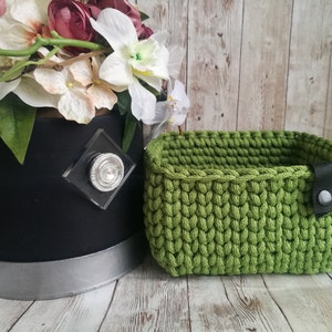 May include: A small, rectangular, lime green crochet basket with a black leather strap and button closure. The basket is next to a black container with a silver rim and a floral arrangement.